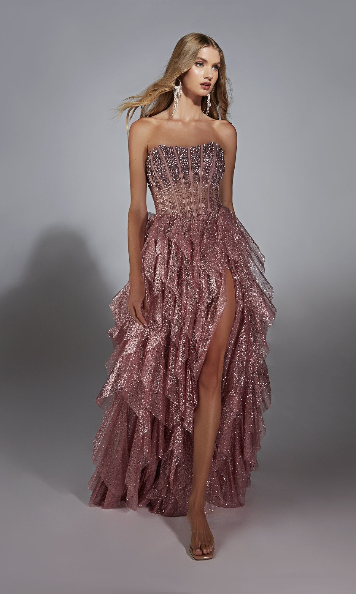 A woman wearing the Alyce 61762 Strapless Long Glitter A-Prom Dress, featuring a sheer bodice, ruffled skirt, and high slit, stands against a plain gray background.
